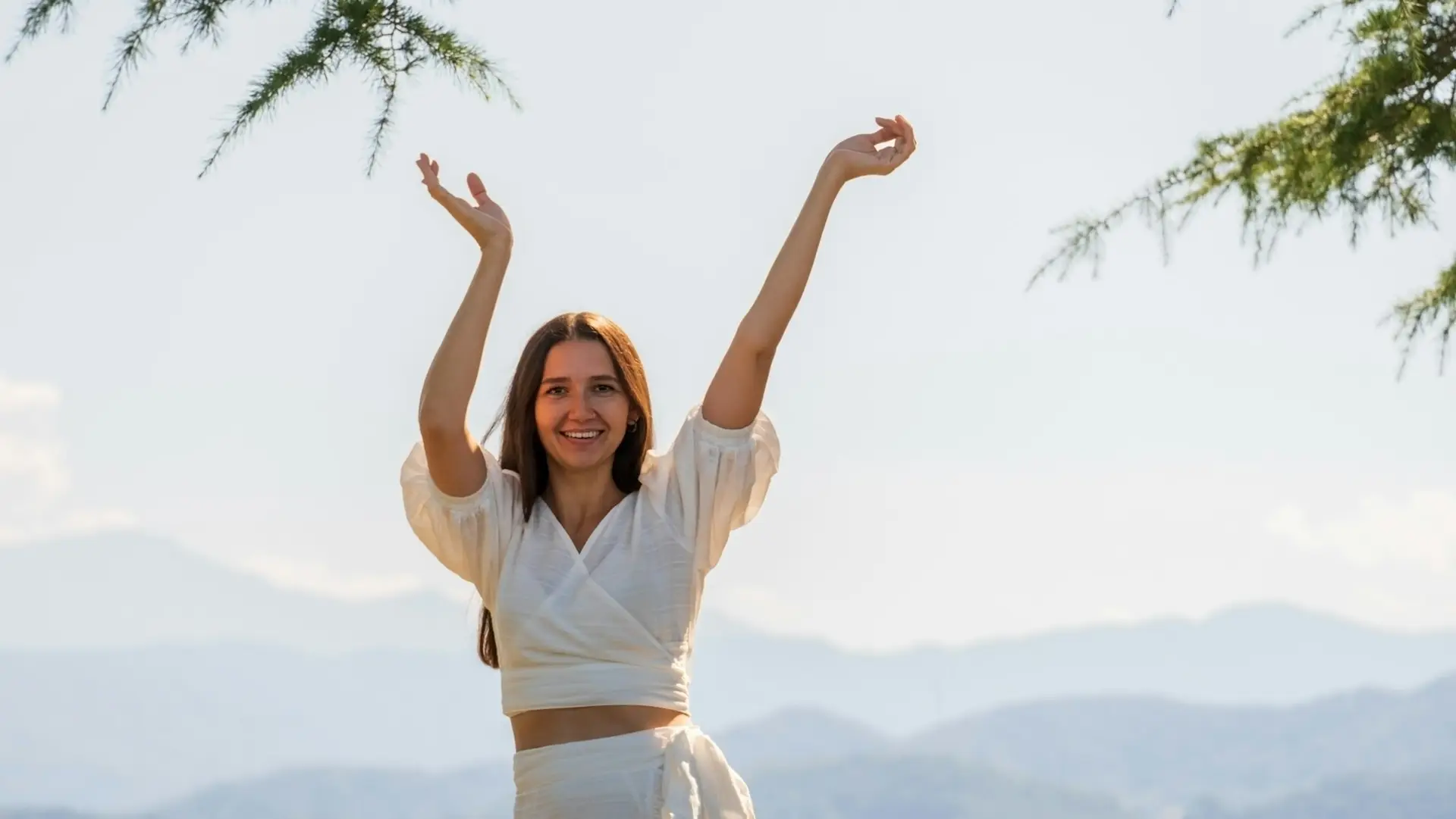A young, beautiful, healthy woman in a white dress is dancing against the background of mountains.